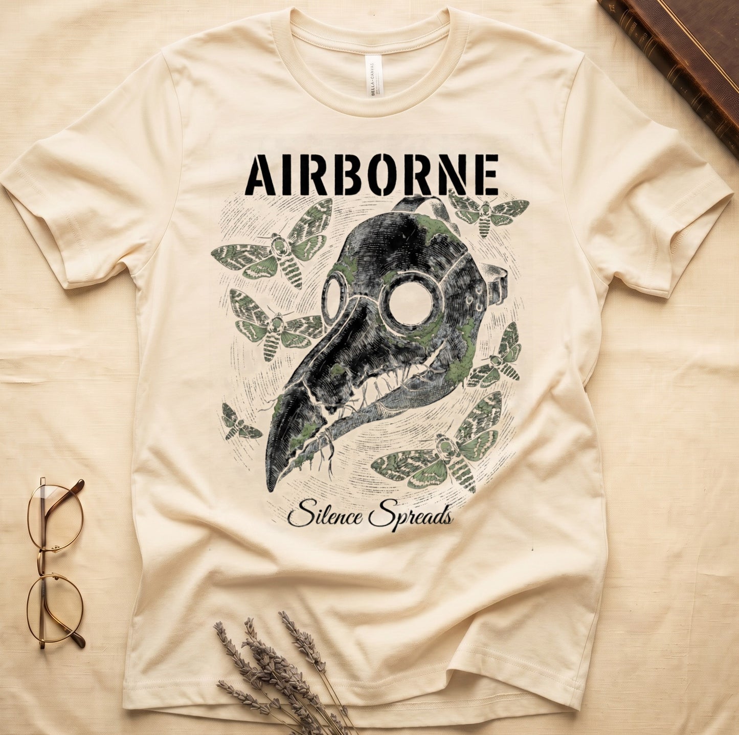 Natural tee with plague doctor mask in cream linework, death's-head moths, "AIRBORNE" top, "Silence Spreads" cursive below.