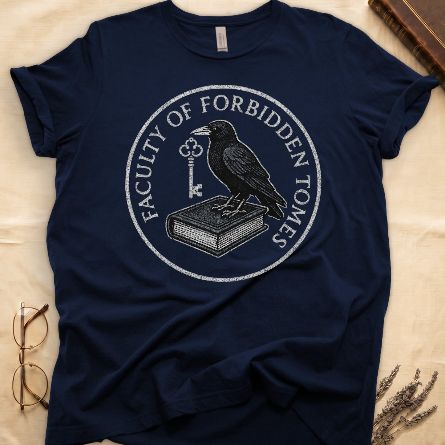 Black tee with circular badge, raven on closed book holding key, "FACULTY OF FORBIDDEN TOMES" curved around in cream.