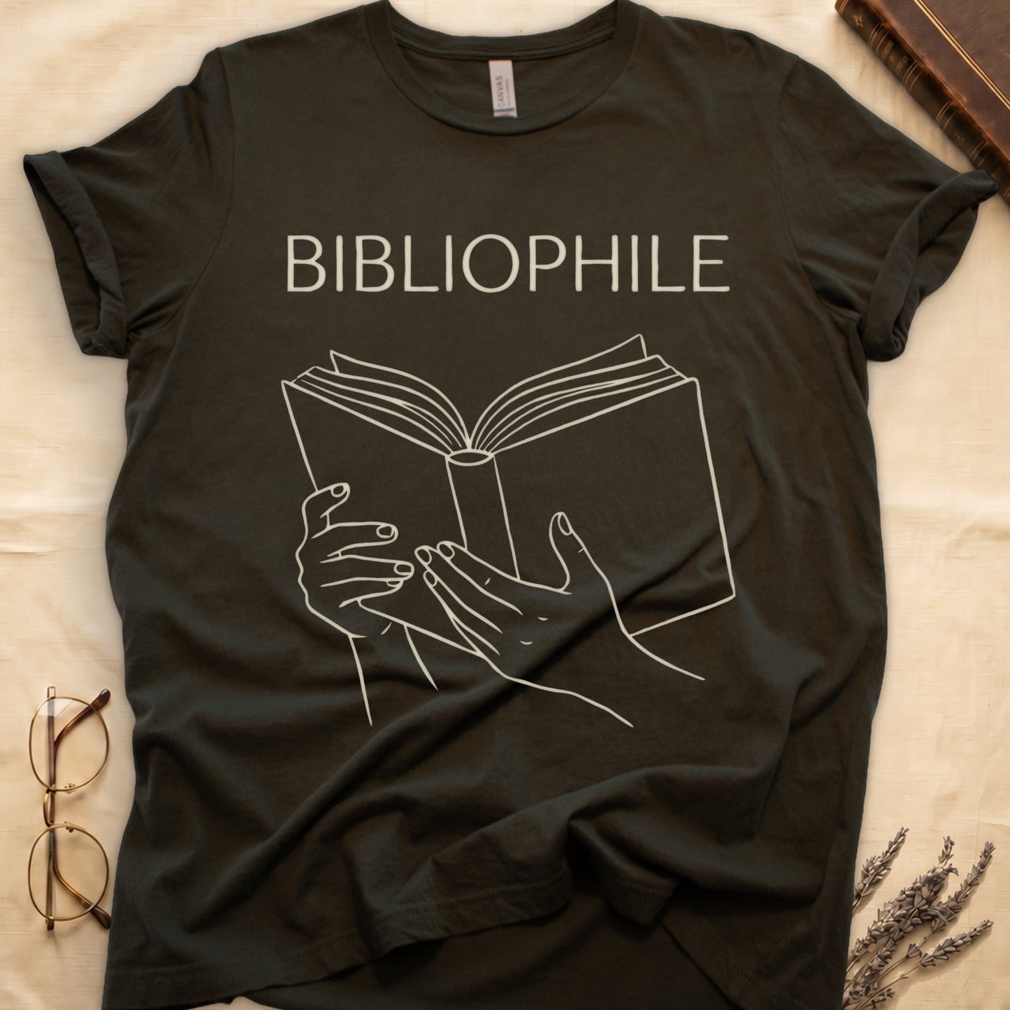 Military Green tee with two hands cradling an open book in cream and brown linework, "BIBLIOPHILE" in serif capitals above.