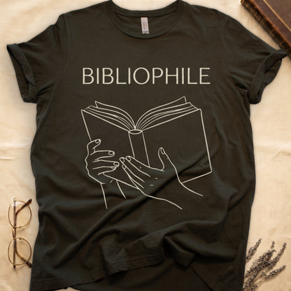 Military Green tee with two hands cradling an open book in cream and brown linework, "BIBLIOPHILE" in serif capitals above.