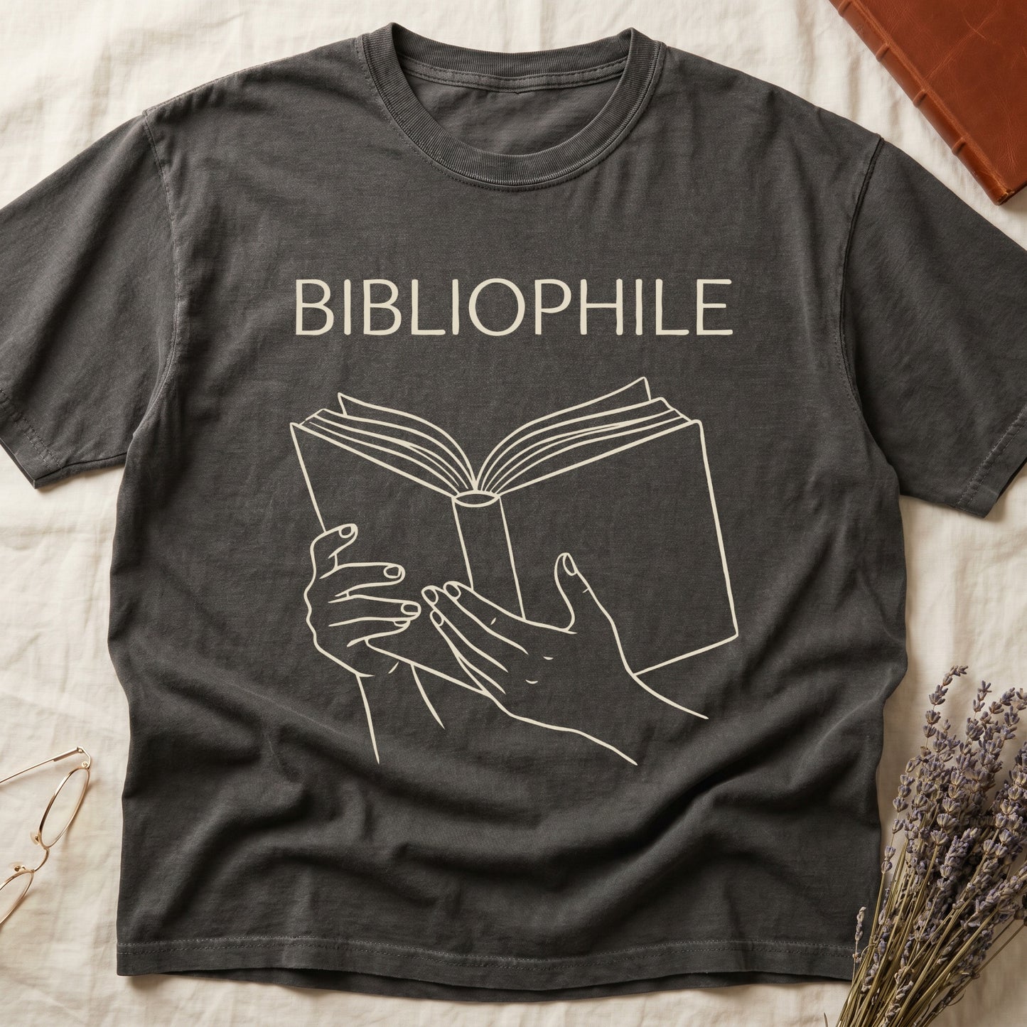 Pepper tee with two hands cradling an open book in cream and brown linework, "BIBLIOPHILE" in serif capitals above.