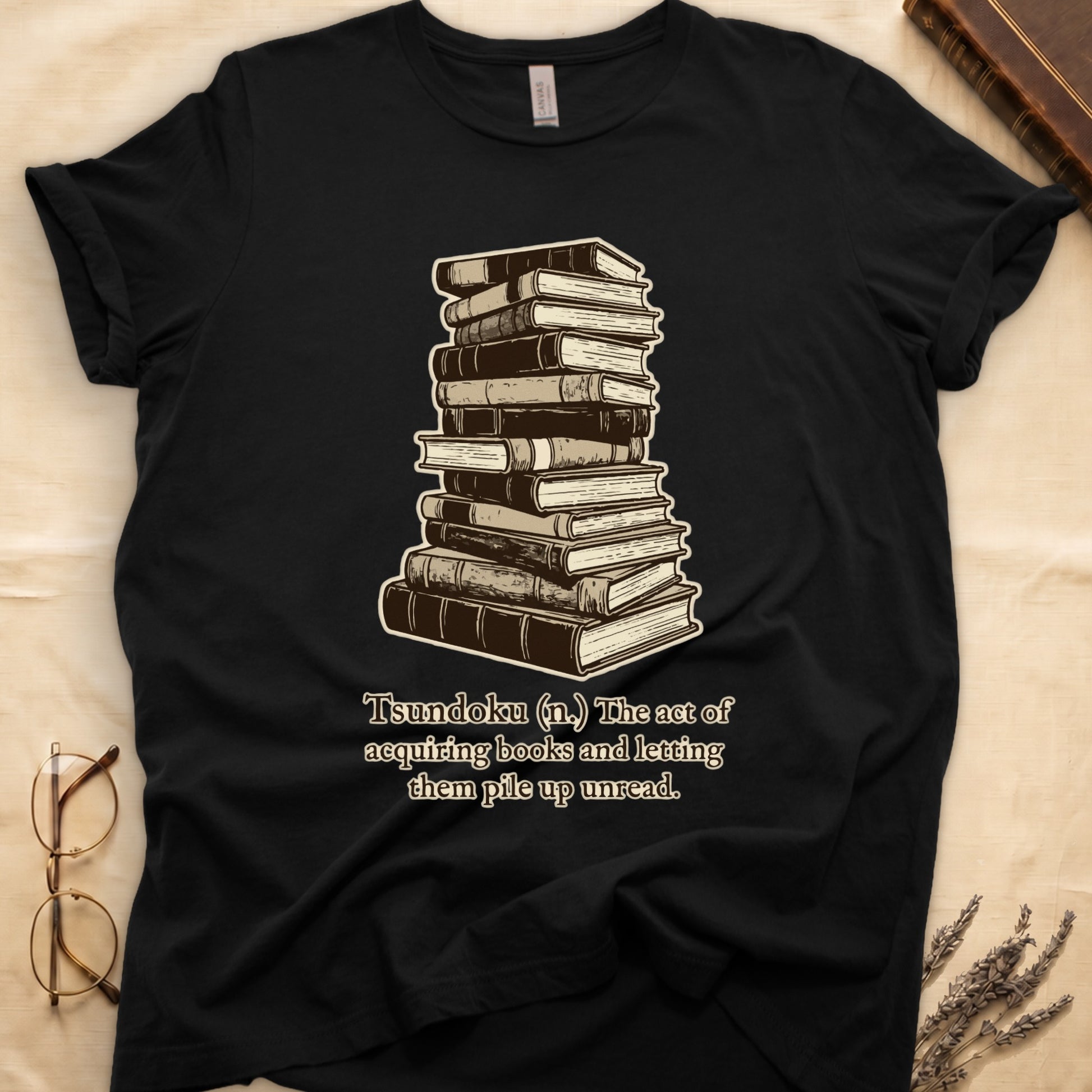 Black tee with stack of vintage hardcovers, "Tsundoku (n.) The act of acquiring books and letting them pile up unread."