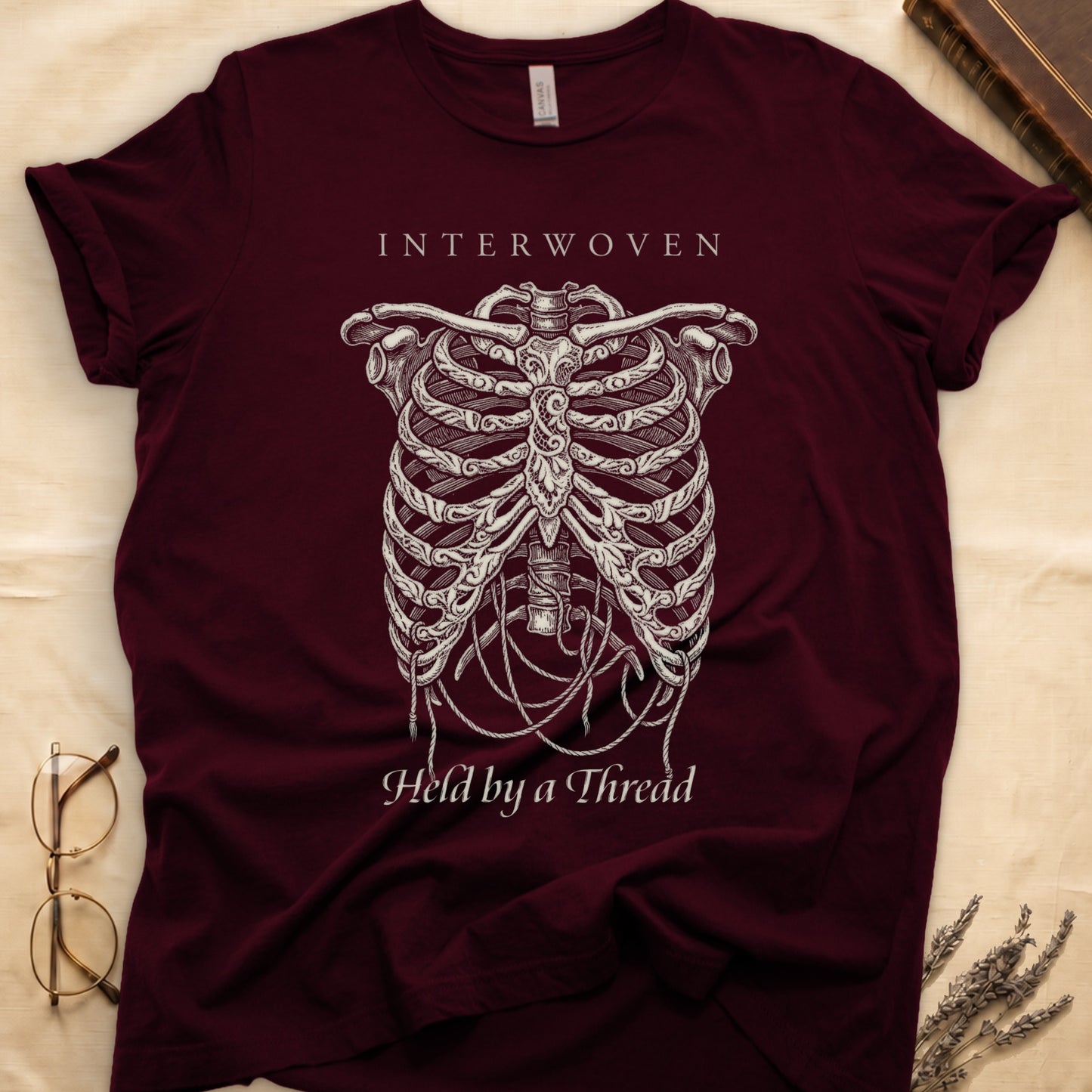 Maroon tee with cream skeletal ribcage suspended by trailing threads, "Held by a Thread" in serif on plate below.