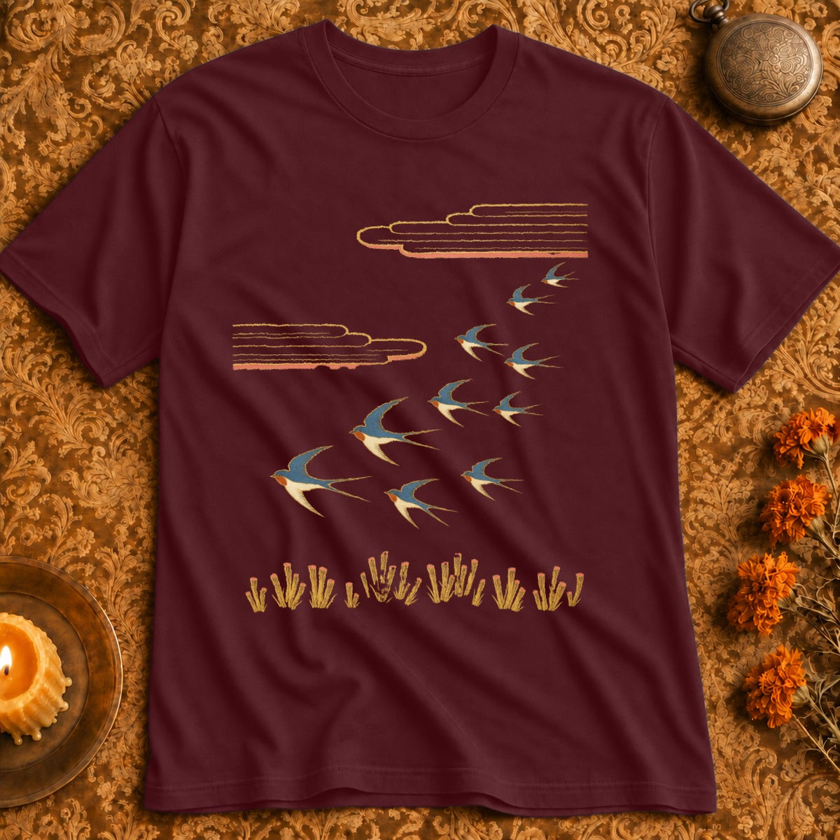Maroon t-shirt with a flock of swallows flying over golden reeds under stylized clouds.