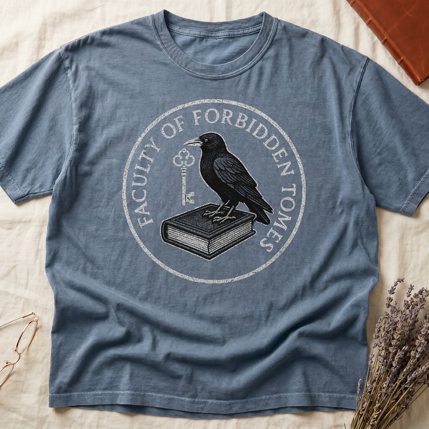 Espresso tee with circular badge, raven on closed book holding key, "FACULTY OF FORBIDDEN TOMES" curved around in cream.