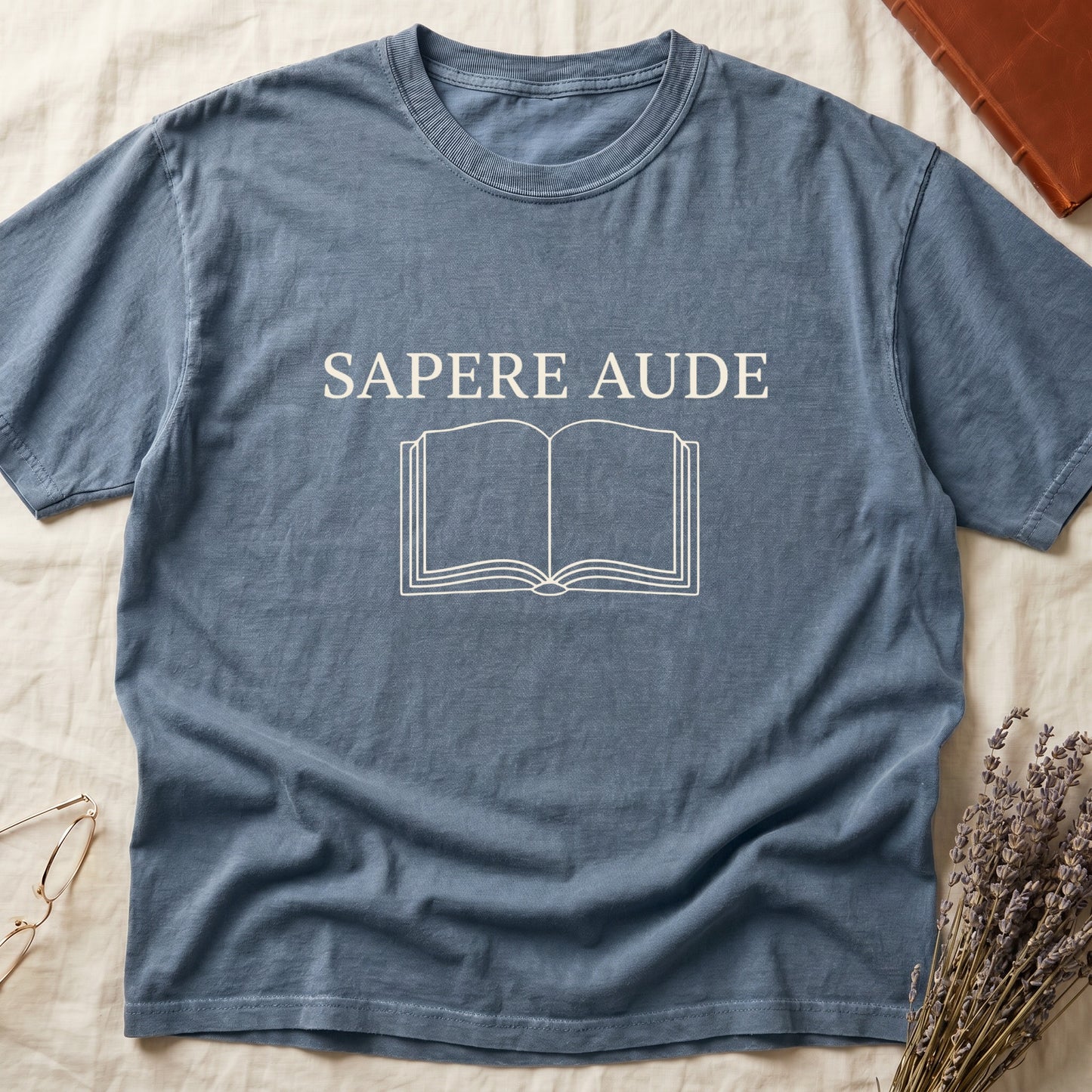 Blue Jean tee with "SAPERE AUDE" in serif capitals above a minimalist open book in cream linework.