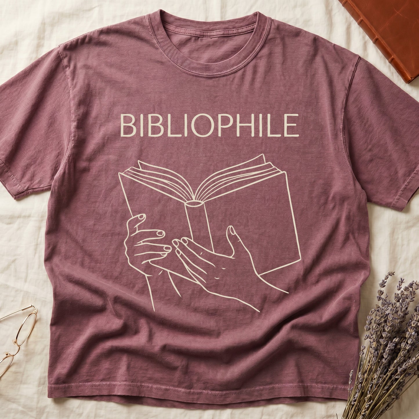 Dark Rose tee with two hands cradling an open book in cream and brown linework, "BIBLIOPHILE" in serif capitals above.