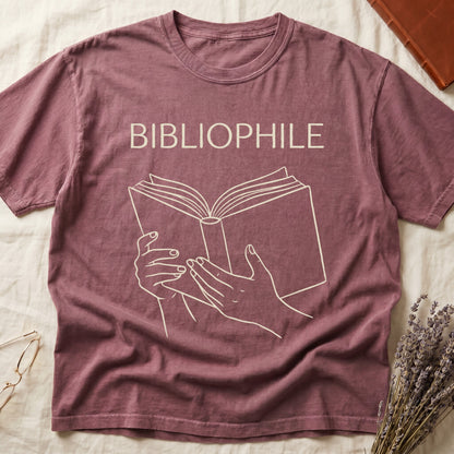 Dark Rose tee with two hands cradling an open book in cream and brown linework, "BIBLIOPHILE" in serif capitals above.