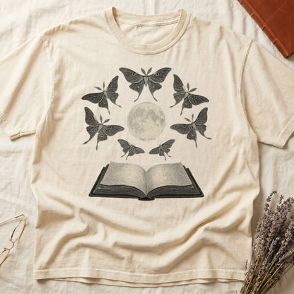 Ivory tee with full moon center, seven luna moths in halo around, open book below, cream-on-black engraving.
