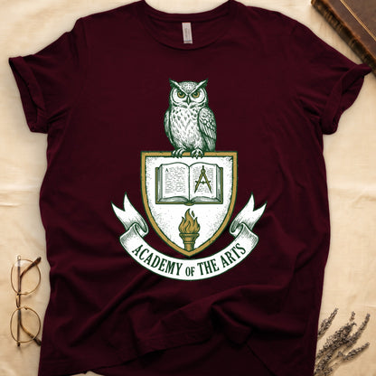 Maroon tee with heraldic shield crest, owl perched on top, open book and dividers center, "ACADEMY OF THE ARTS" on a banner.