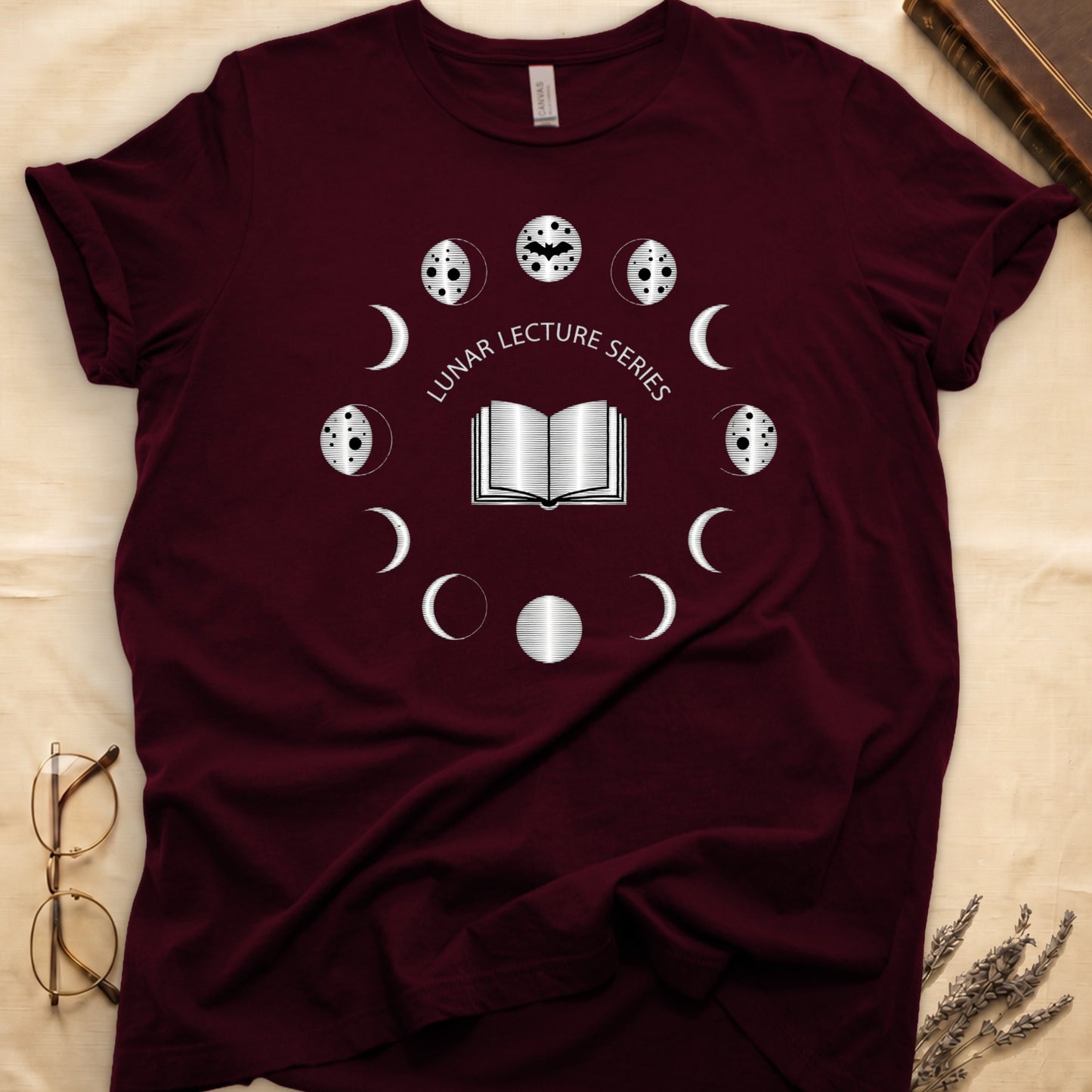 Maroon tee with open book at center, ring of cream moon phases around, curved "LUNAR LECTURE SERIES" double-arc above.