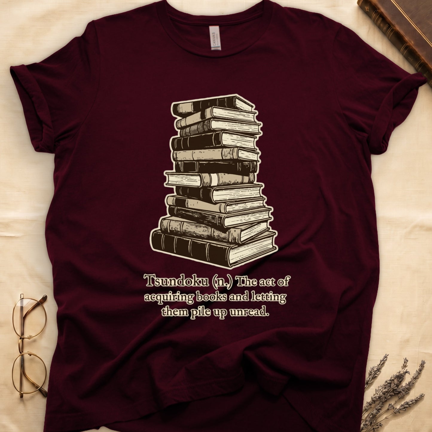 Maroon tee with stack of vintage hardcovers, "Tsundoku (n.) The act of acquiring books and letting them pile up unread."