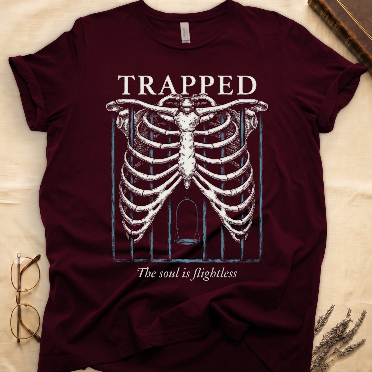Maroon tee with anatomical ribcage on prison bars, bird swing inside, "TRAPPED" top, "The soul is flightless" italic below.