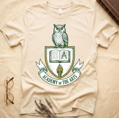 Natural tee with heraldic shield crest, owl perched on top, open book and dividers center, "ACADEMY OF THE ARTS" on a banner.