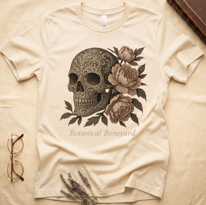 Natural tee with filigree-engraved skull on left, peach-cream peonies on right, crossed bones below, "Botanical Boneyard."