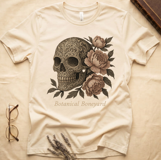 Natural tee with filigree-engraved skull on left, peach-cream peonies on right, crossed bones below, "Botanical Boneyard."