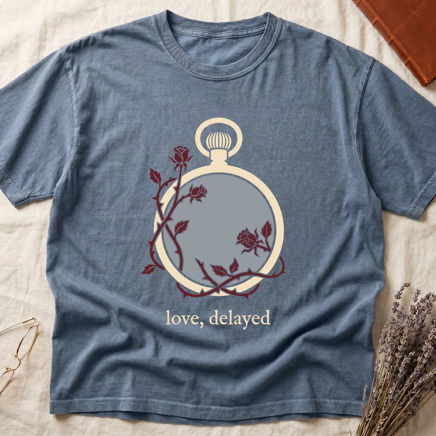 Blue Jean tee with vintage cream pocket watch and dark red roses entwined, "love, delayed" in cream serif below.