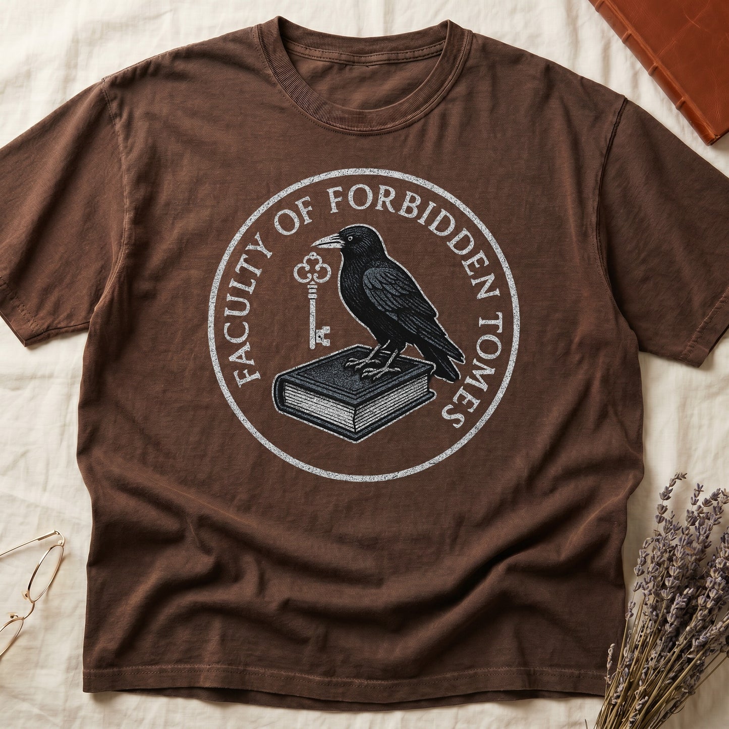 Black tee with circular badge, raven on closed book holding key, "FACULTY OF FORBIDDEN TOMES" curved around in cream.