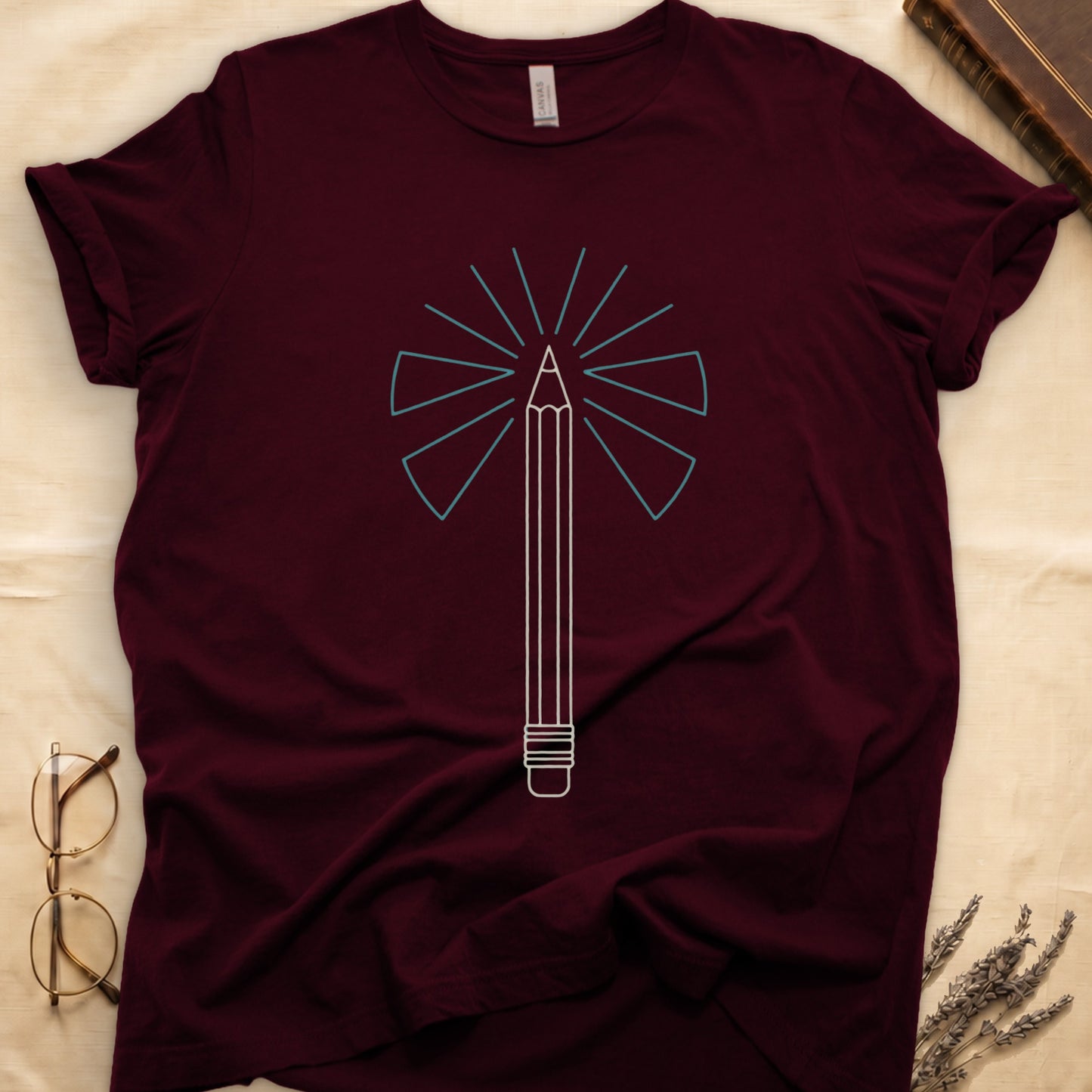 Maroon tee with single vertical graphite pencil in cream linework, fan of teal and grey rays radiating behind.
