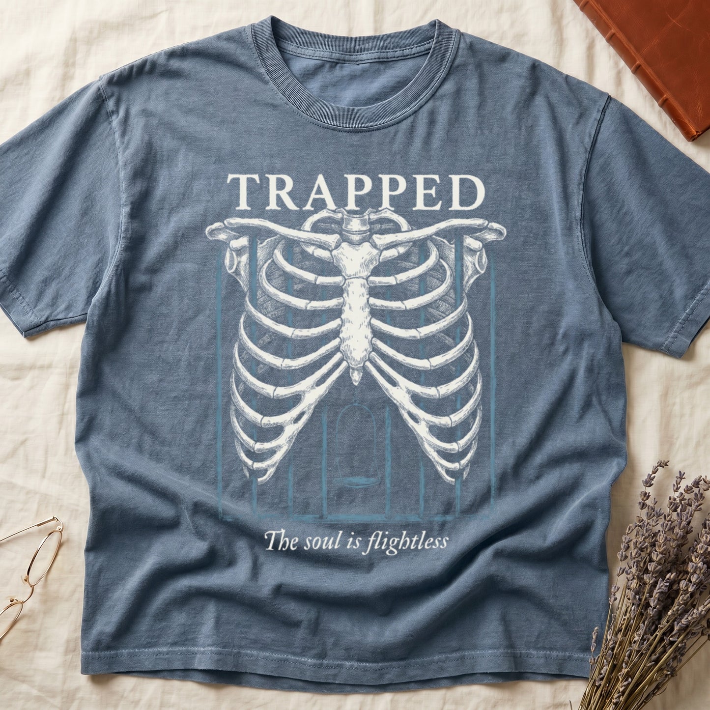 Blue Jean tee with anatomical ribcage on prison bars, bird swing inside, "TRAPPED" top, "The soul is flightless" italic below.