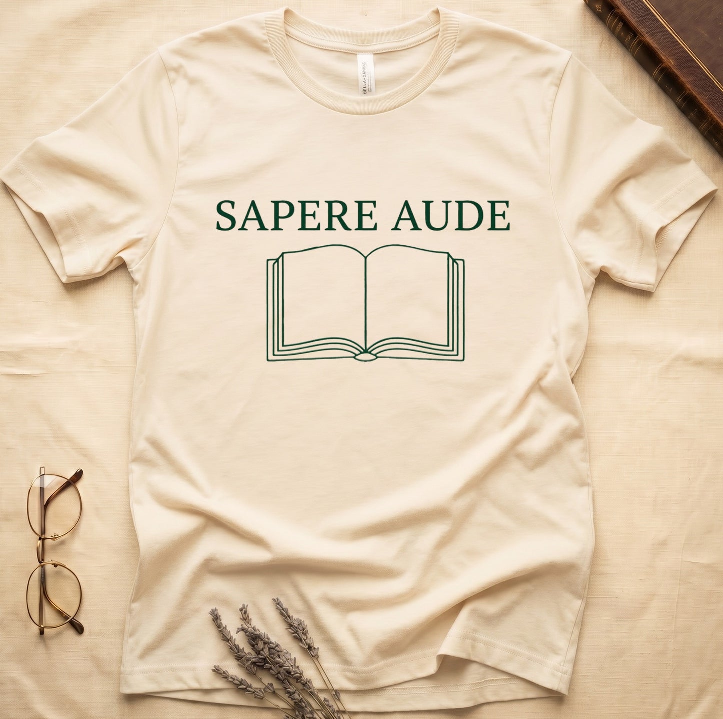 Natural tee with "SAPERE AUDE" in serif capitals above a minimalist open book in cream linework.