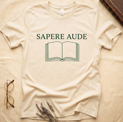 Natural tee with "SAPERE AUDE" in serif capitals above a minimalist open book in cream linework.