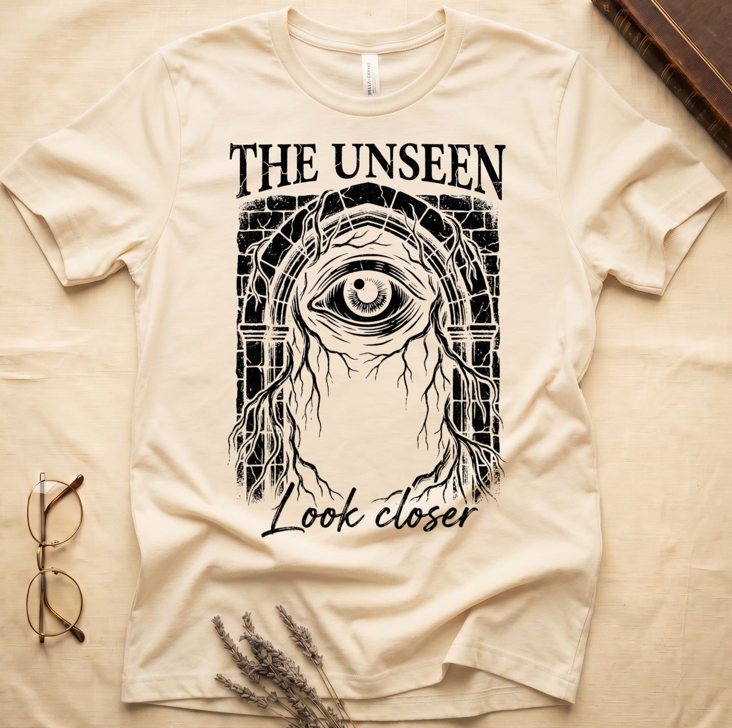 Natural tee with Gothic stone arch, open eye, roots trailing, "THE UNSEEN" top, "Look closer" cursive below.