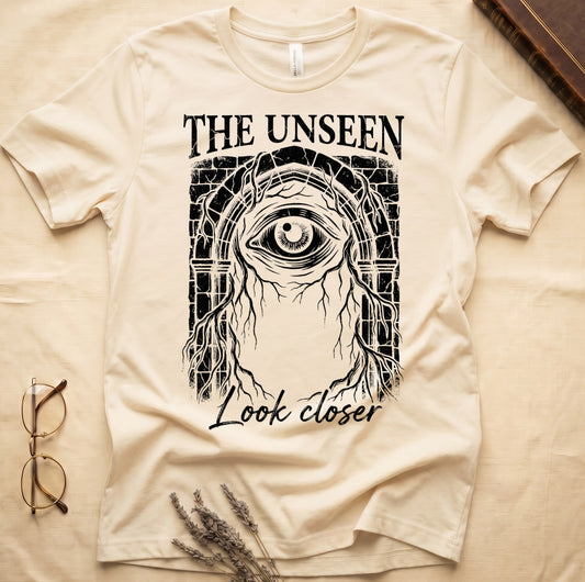 Natural tee with Gothic stone arch, open eye, roots trailing, "THE UNSEEN" top, "Look closer" cursive below.