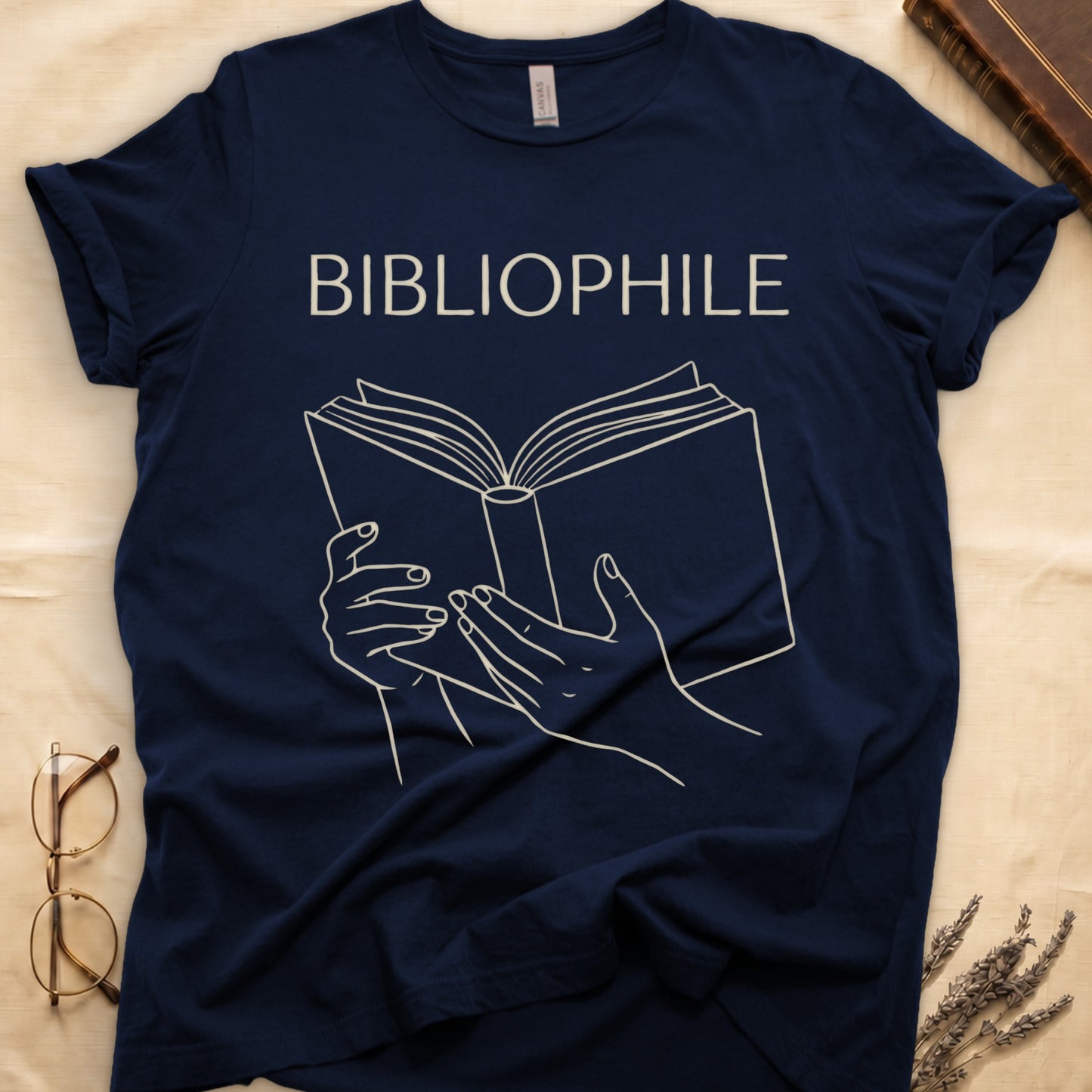 Navy tee with two hands cradling an open book in cream and brown linework, "BIBLIOPHILE" in serif capitals above.
