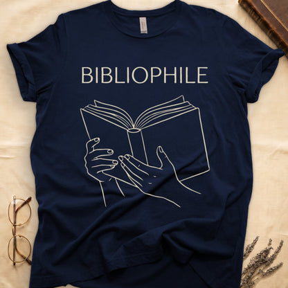 Navy tee with two hands cradling an open book in cream and brown linework, "BIBLIOPHILE" in serif capitals above.