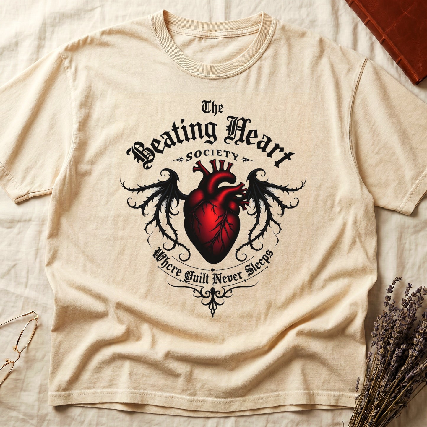 Ivory tee with anatomical red heart, gothic wings, "The Beating Heart SOCIETY," "Where Guilt Never Sleeps" below.