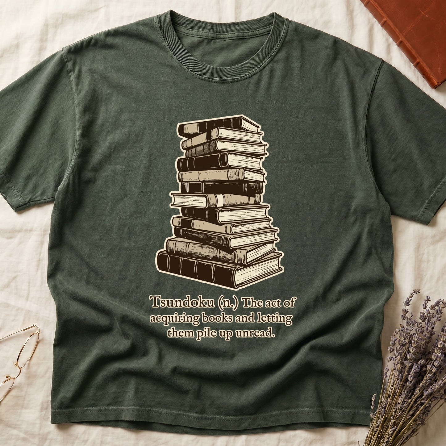 Green tee with stack of vintage hardcovers, "Tsundoku (n.) The act of acquiring books and letting them pile up unread."