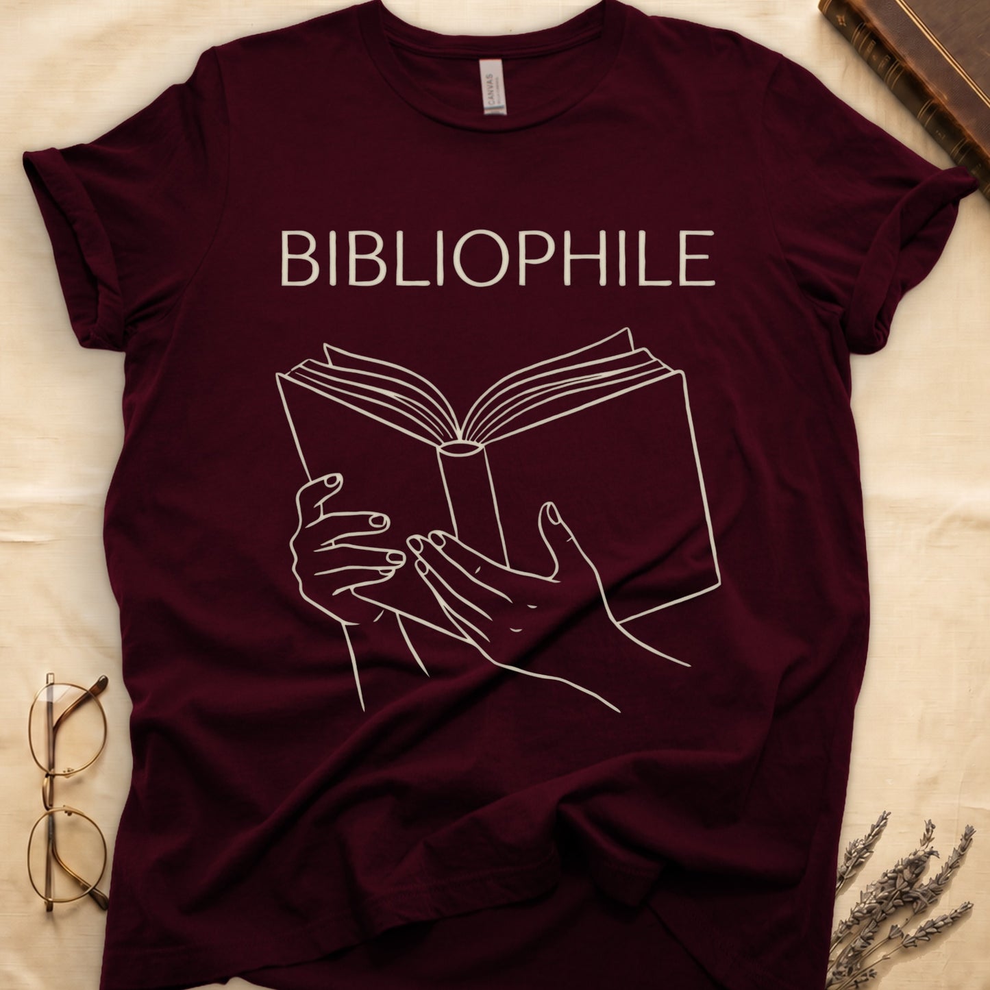 Maroon tee with two hands cradling an open book in cream and brown linework, "BIBLIOPHILE" in serif capitals above.