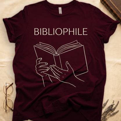 Maroon tee with two hands cradling an open book in cream and brown linework, "BIBLIOPHILE" in serif capitals above.