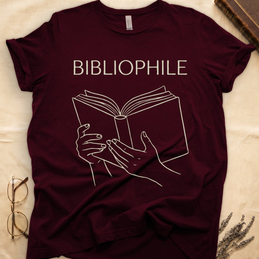 Maroon tee with two hands cradling an open book in cream and brown linework, "BIBLIOPHILE" in serif capitals above.