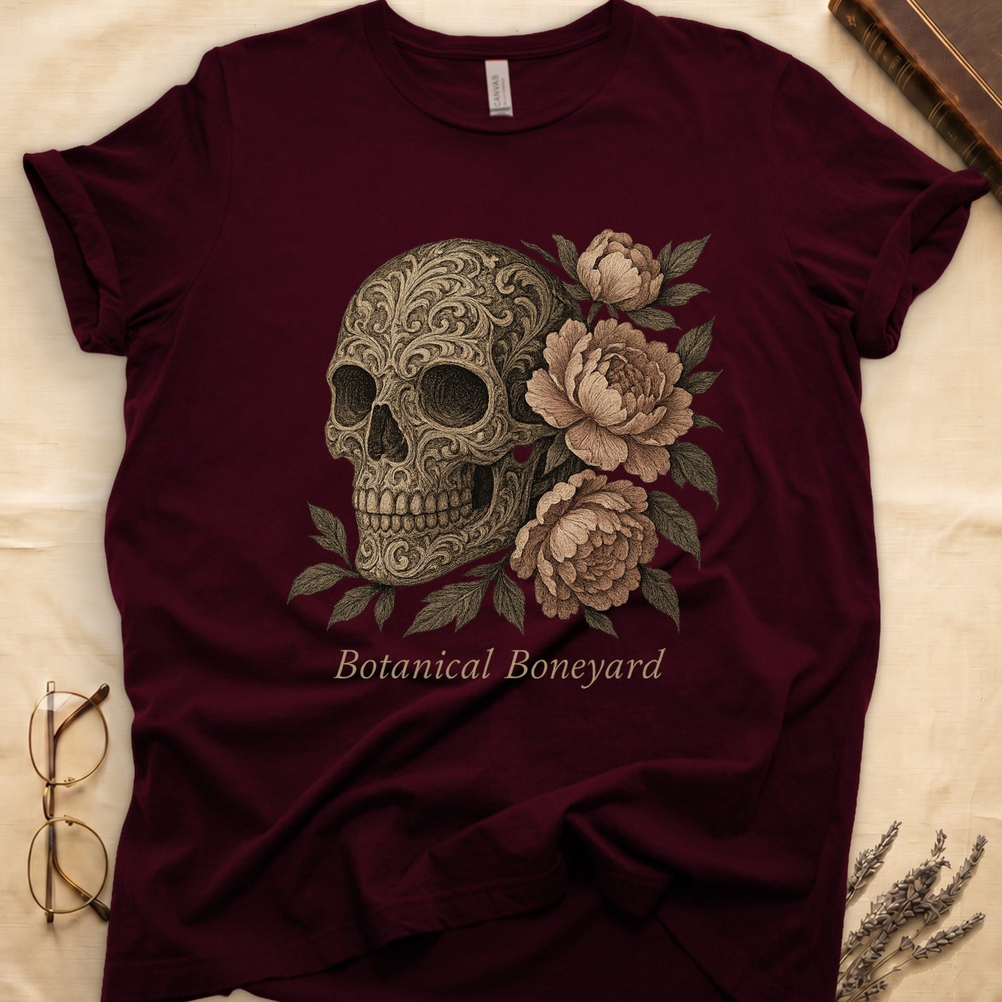 Maroon tee with filigree-engraved skull on left, peach-cream peonies on right, crossed bones below, "Botanical Boneyard."