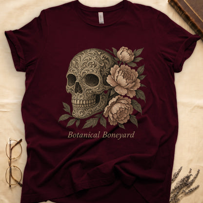Maroon tee with filigree-engraved skull on left, peach-cream peonies on right, crossed bones below, "Botanical Boneyard."