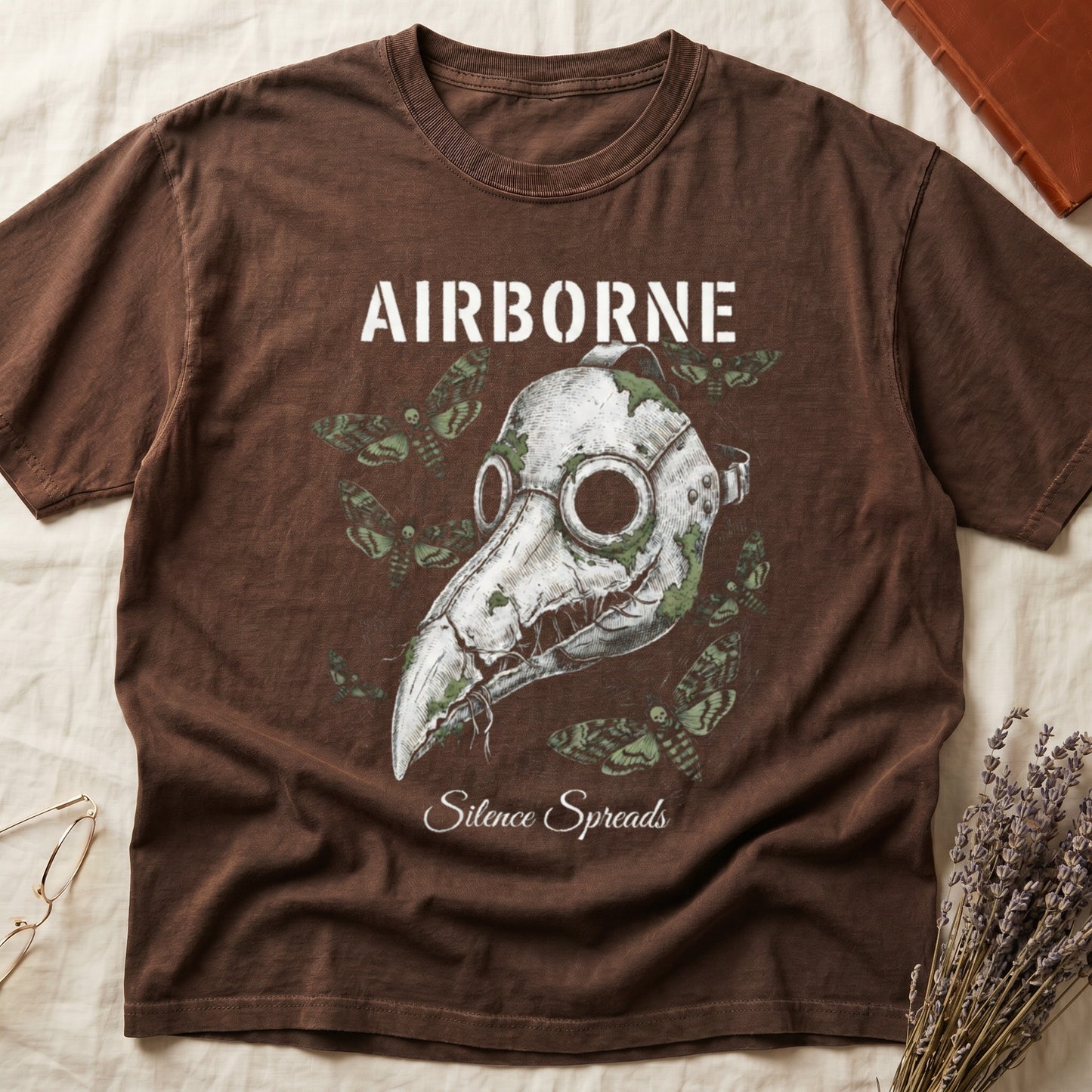 Espresso tee with plague doctor mask in cream linework, death's-head moths, "AIRBORNE" top, "Silence Spreads" cursive below.