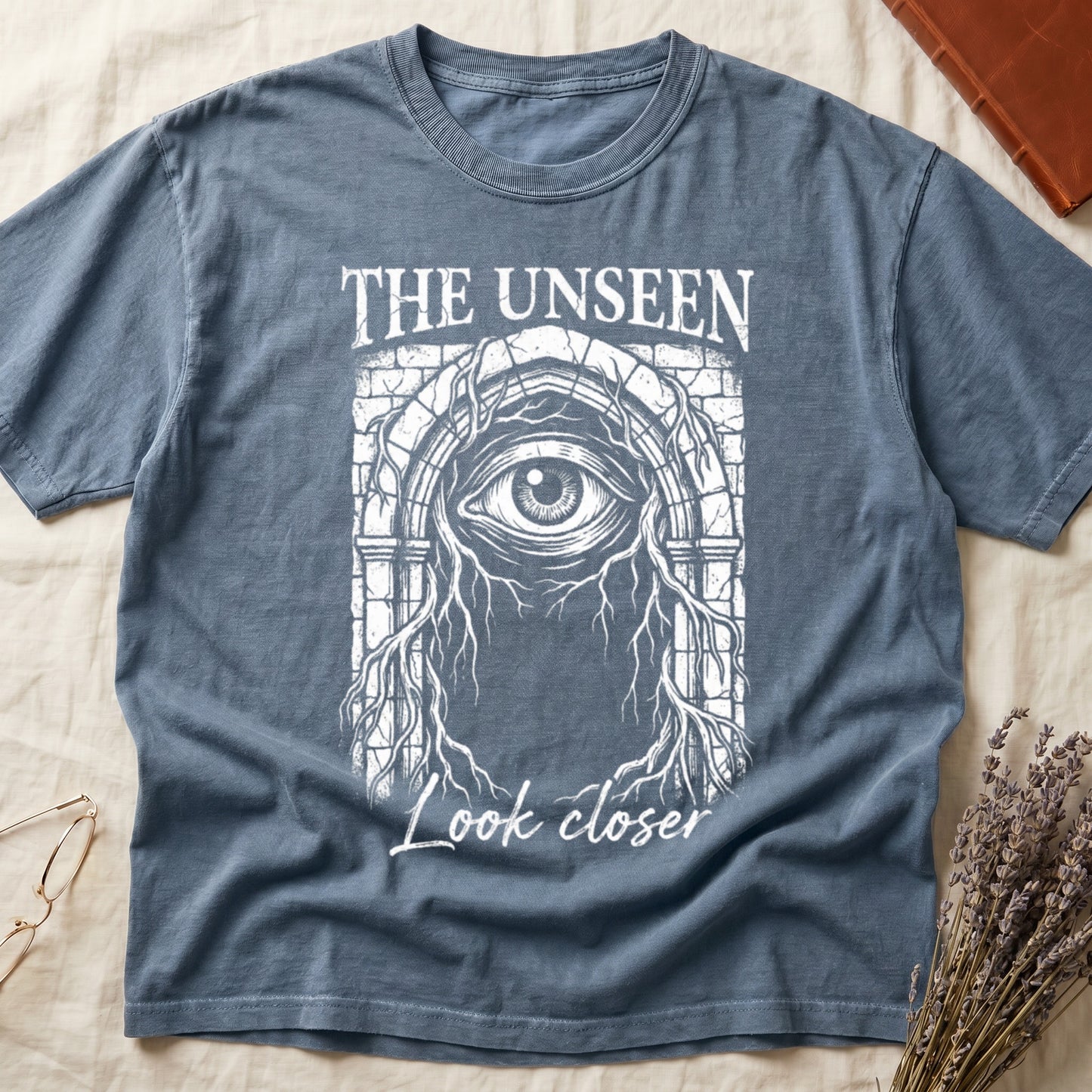 Blue Jean tee with Gothic stone arch, open eye, roots trailing, "THE UNSEEN" top, "Look closer" cursive below.