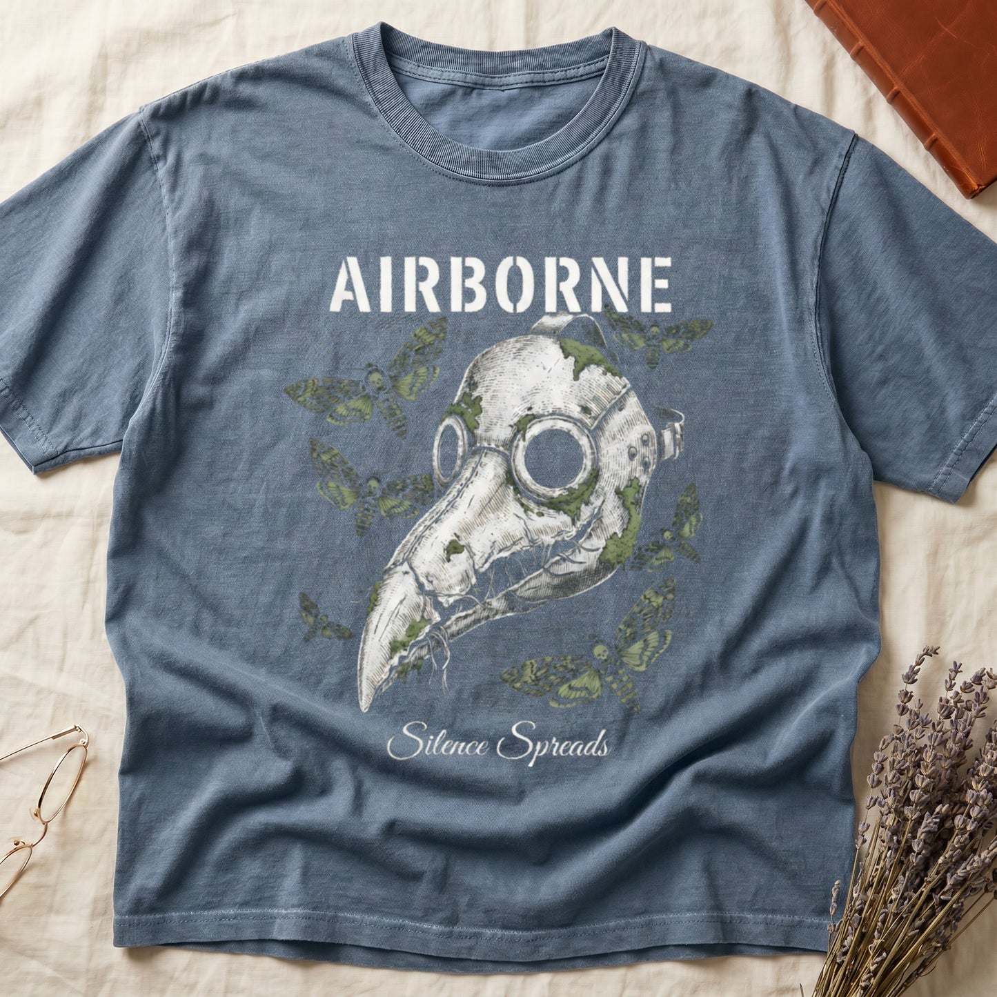Blue Jean tee with plague doctor mask in cream linework, death's-head moths, "AIRBORNE" top, "Silence Spreads" cursive below.