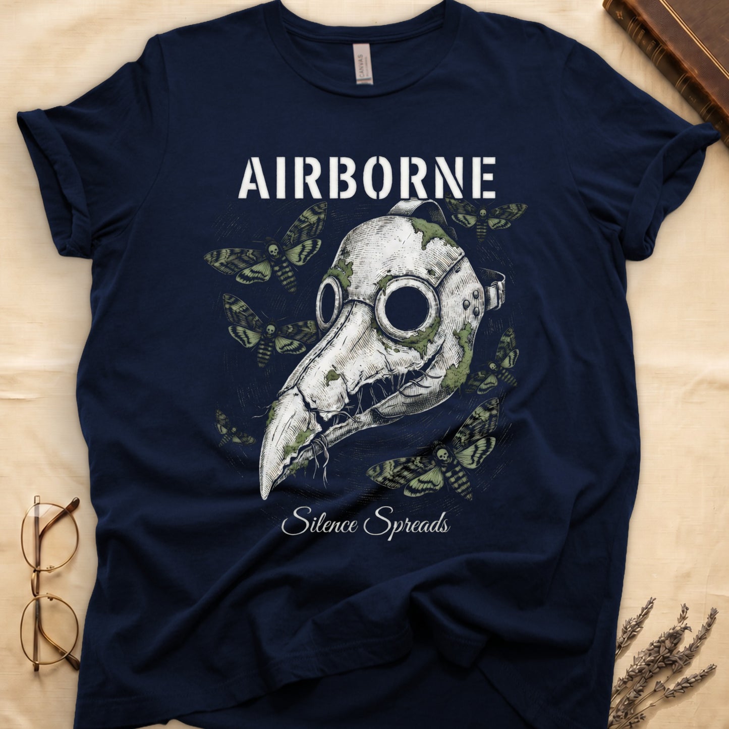 Navy tee with plague doctor mask in cream linework, death's-head moths, "AIRBORNE" top, "Silence Spreads" cursive below.