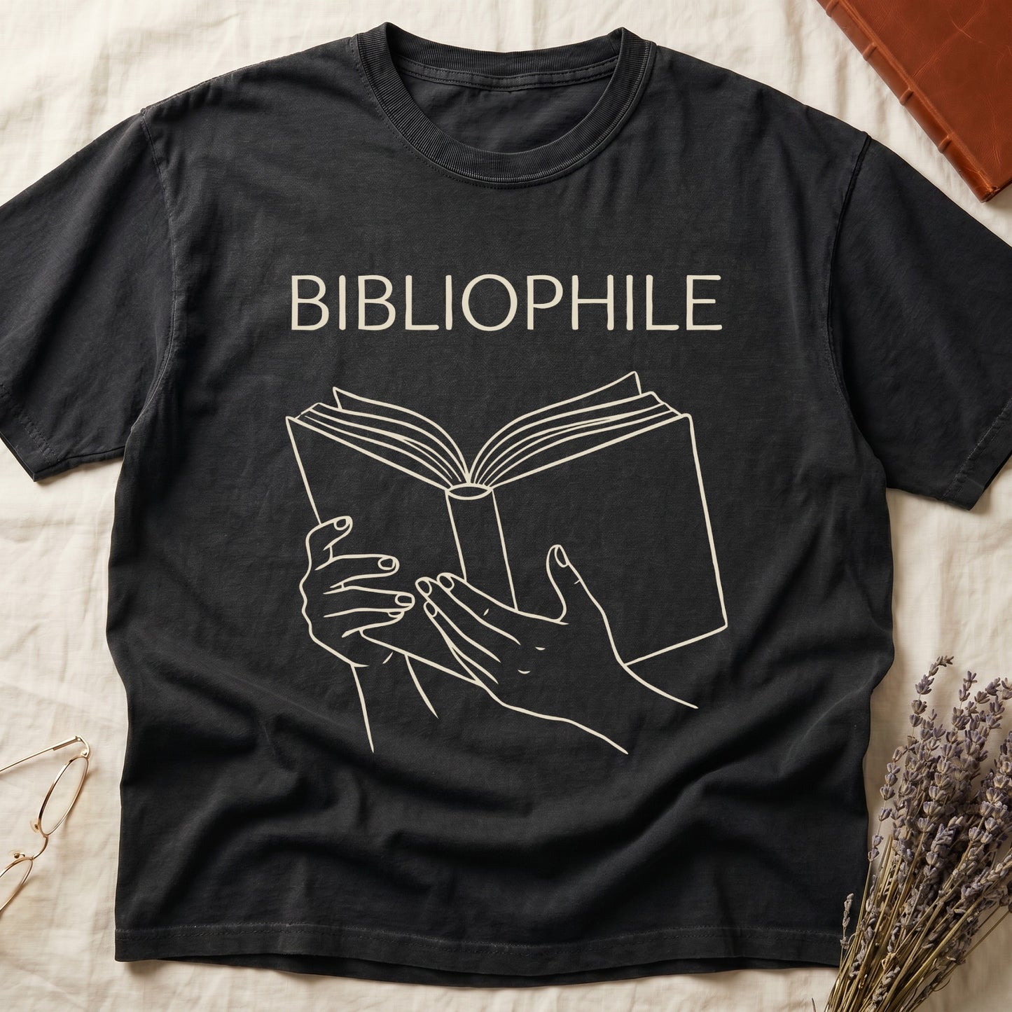 Black tee with two hands cradling an open book in cream and brown linework, "BIBLIOPHILE" in serif capitals above.