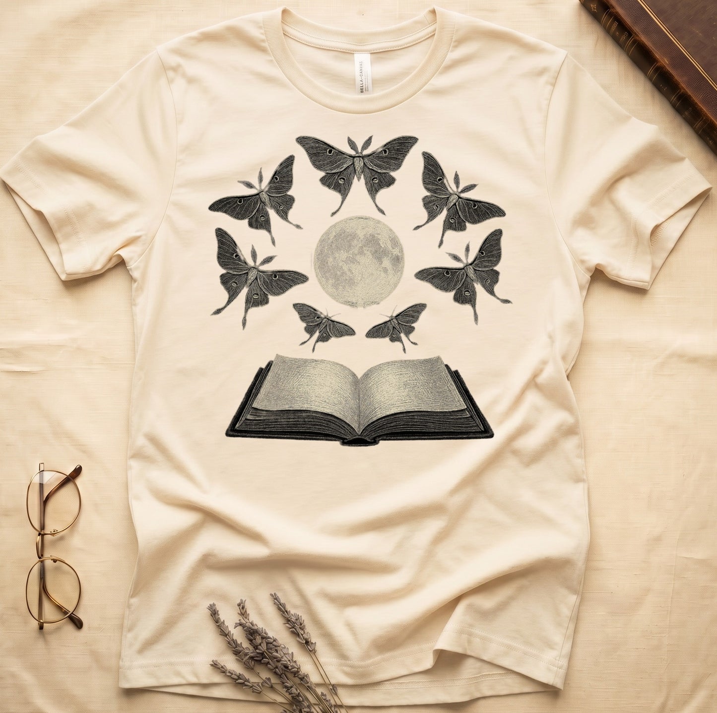Natural tee with full moon center, seven luna moths in halo around, open book below, cream-on-black engraving.