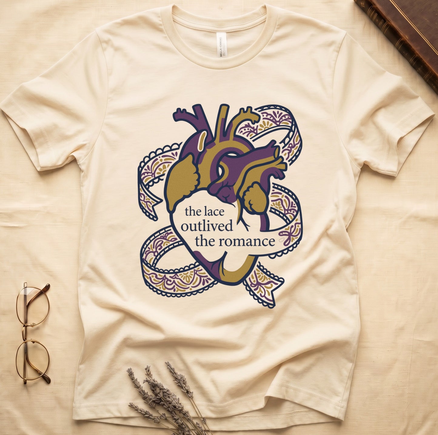Natural tee with anatomical heart in purple and gold wrapped in lace ribbons, "the lace outlived the romance" in serif.