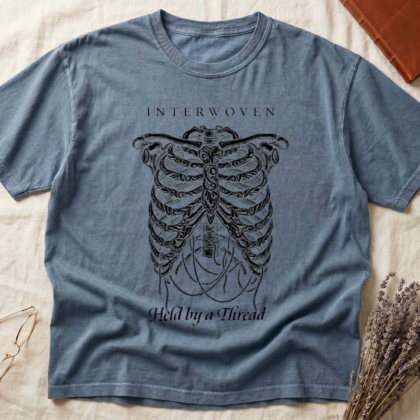 Blue Jean tee with cream skeletal ribcage suspended by trailing threads, "Held by a Thread" in serif on plate below.