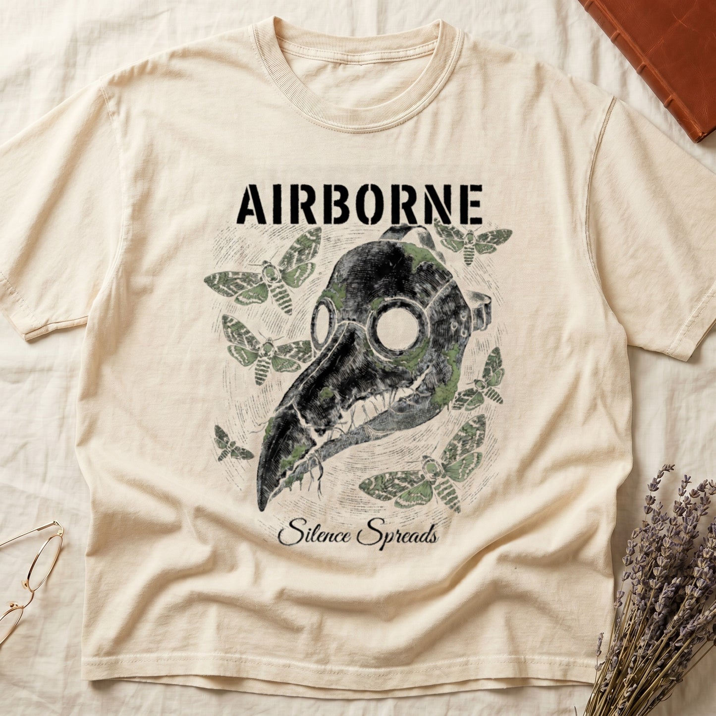 Ivory tee with plague doctor mask in cream linework, death's-head moths, "AIRBORNE" top, "Silence Spreads" cursive below.