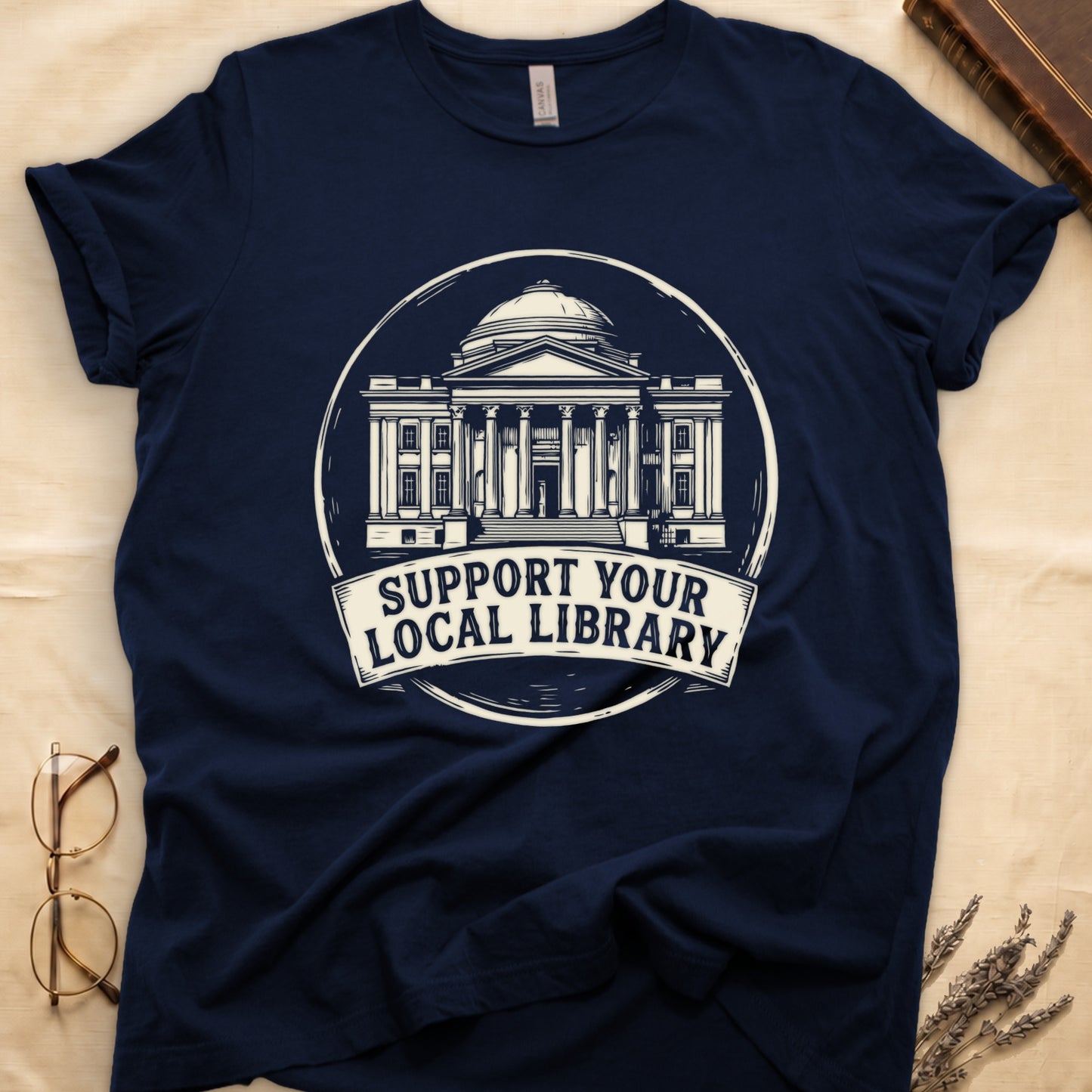 Navy tee with circular cream badge of a classical library building, "SUPPORT YOUR LOCAL LIBRARY" on a banner below.