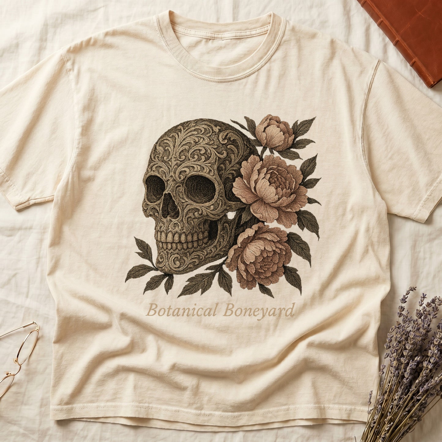 Ivory tee with filigree-engraved skull on left, peach-cream peonies on right, crossed bones below, "Botanical Boneyard."