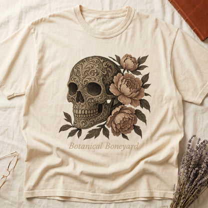 Ivory tee with filigree-engraved skull on left, peach-cream peonies on right, crossed bones below, "Botanical Boneyard."