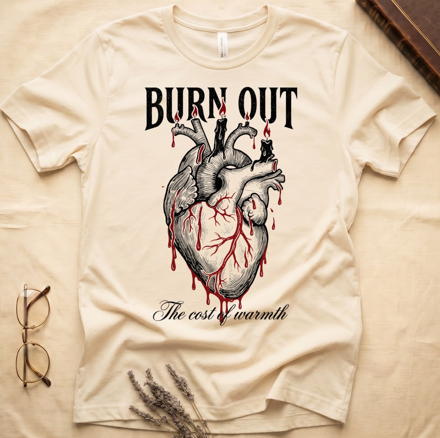 Natural tee with anatomical heart and melted candles dripping red wax, "BURN OUT" top, "The cost of warmth" cursive below.